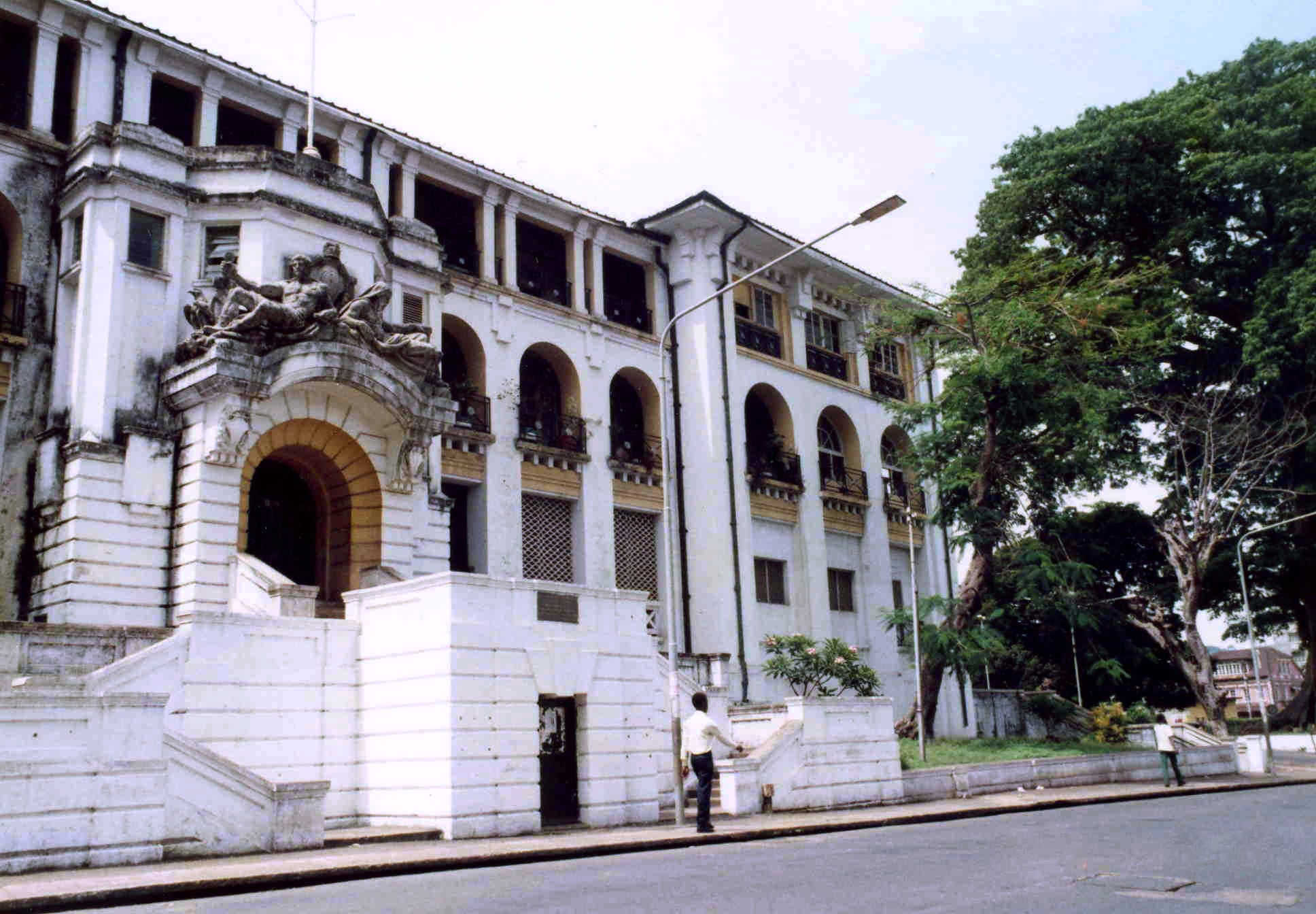 The white colonnaded Law Courts building in central Freetown, with an arched entrance and a large tree at the right edge of the frame.